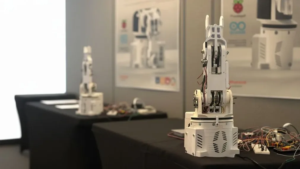 Bournemouth University case study · two 3D-printed white robot arms with visible mechanical detail and Raspberry Pi / Arduino electronics, on expo tables